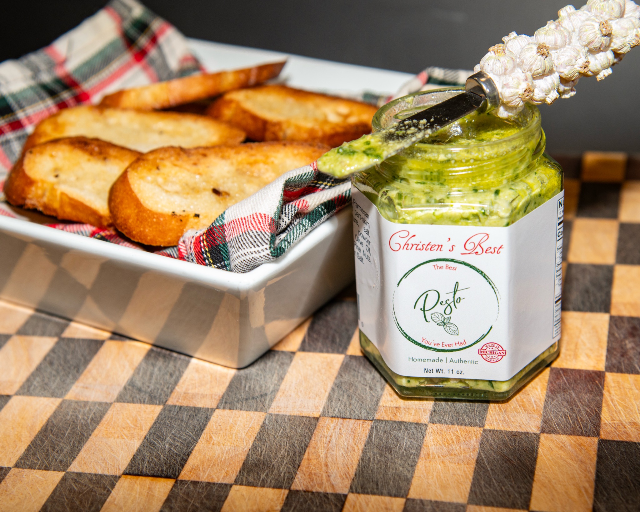 A jar of Christen's Best pesto with a garlic-handled spreader, next to a dish of toasted crostini in a plaid napkin