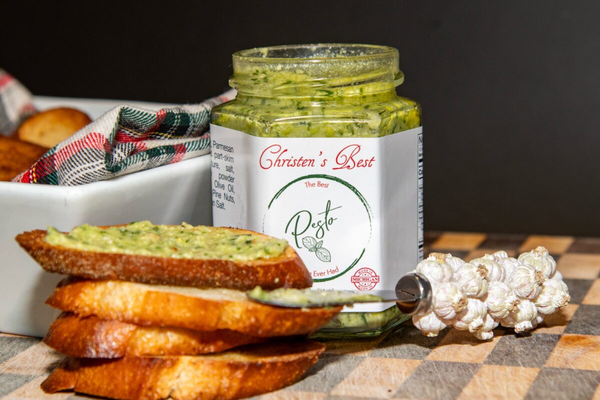 A stack of toasted baguette slices, the top one spread with basil pesto, in front of an open jar