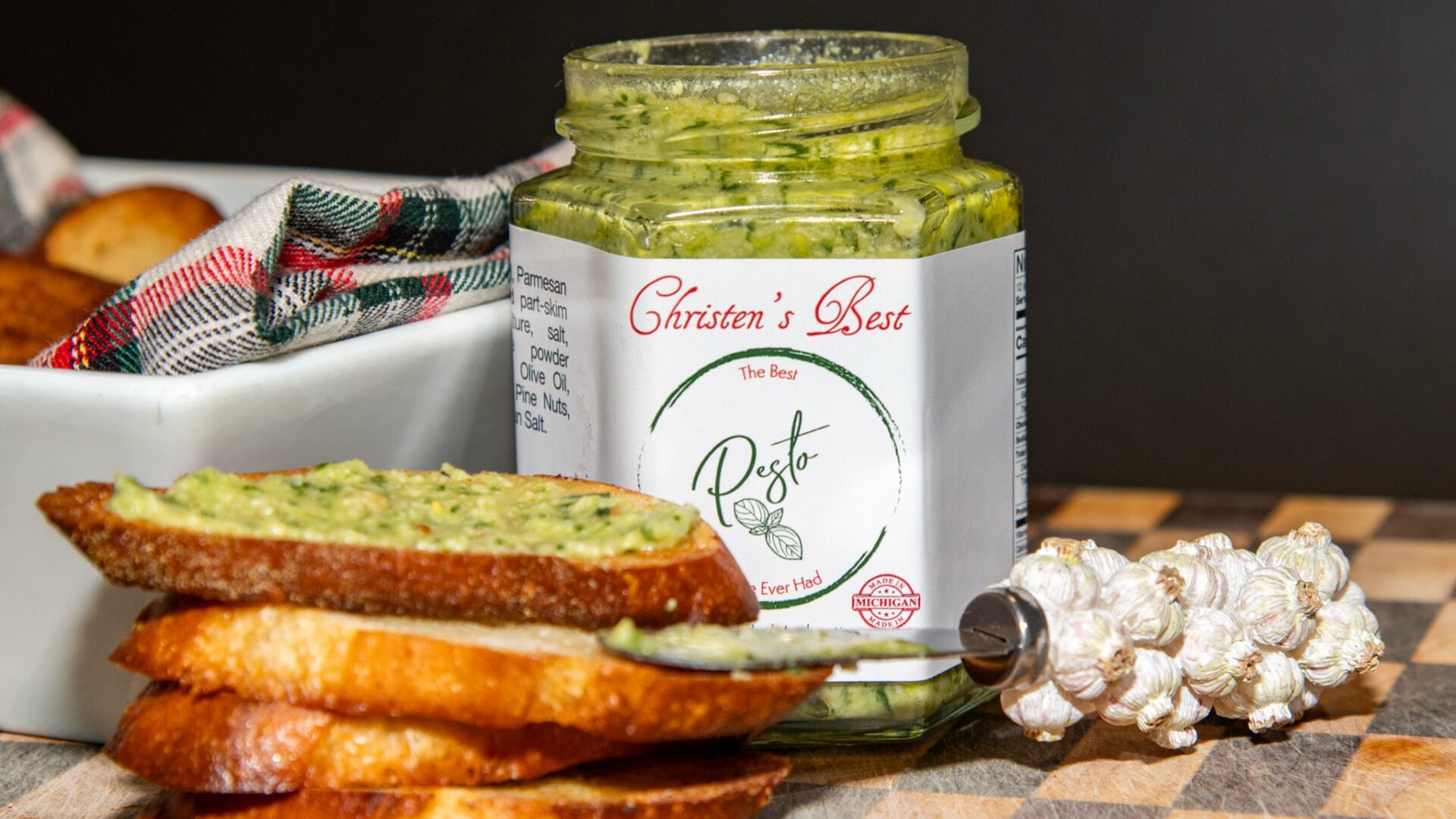 A stack of toasted baguette slices, the top one spread with basil pesto, in front of an open jar