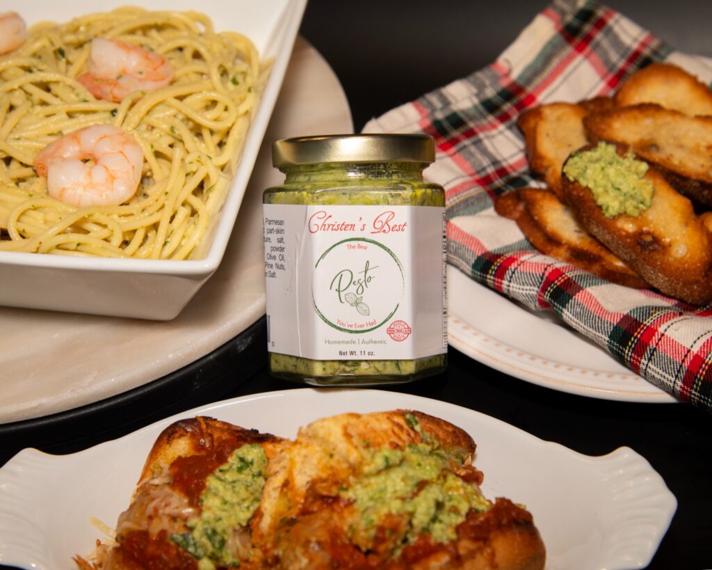 A jar of Christen's Best pesto surrounded by shrimp spaghetti, pesto crostini and a toasted sub