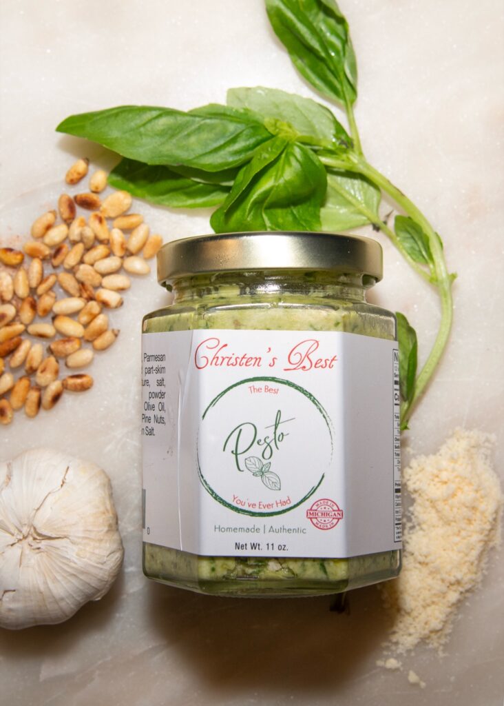 A jar of Christen's Best pesto on marble, surrounded by fresh basil, pine nuts, a head of garlic and grated Parmigiano