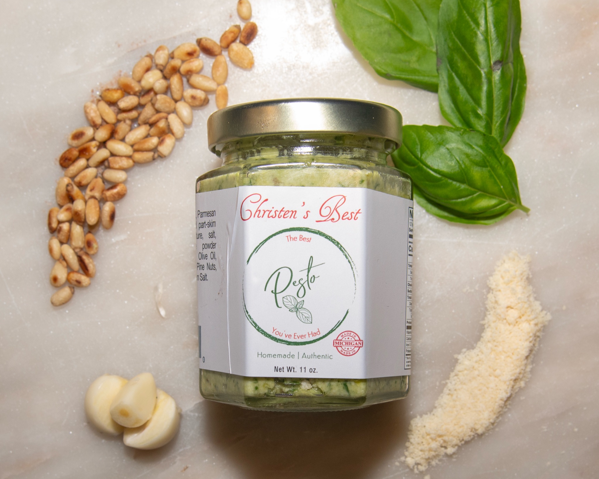 A jar of Christen's Best pesto on marble with basil leaves, pine nuts, garlic cloves and grated Parmigiano