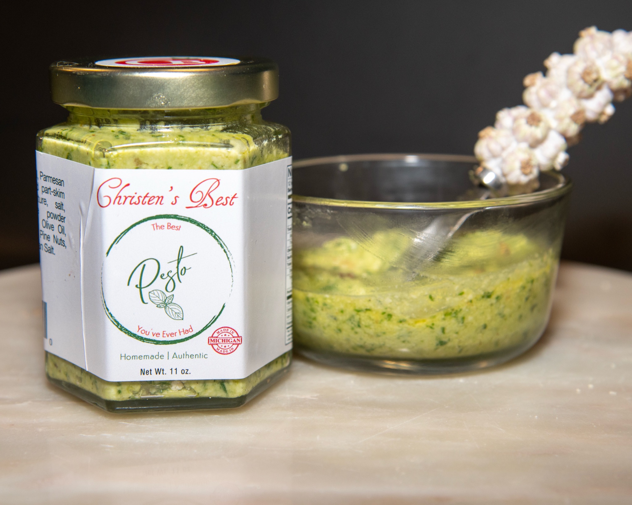 A jar of Christen's Best pesto next to a glass bowl of pesto with a garlic-handled spoon