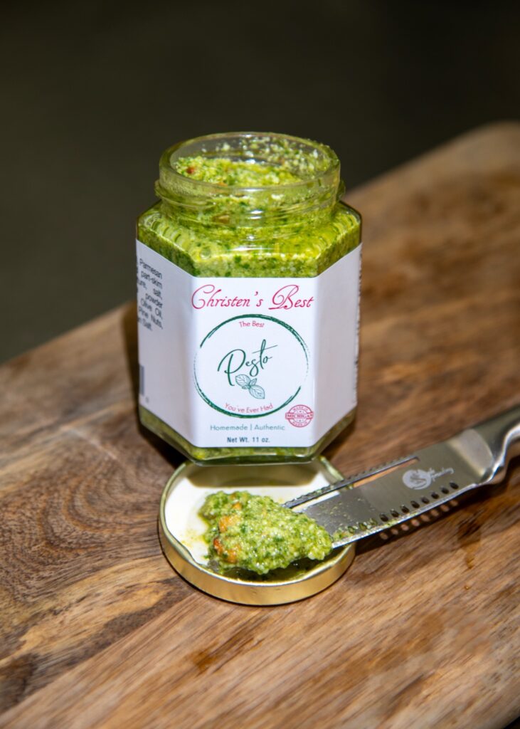 An open jar of basil pesto on a cutting board, with a spreader resting on the lid