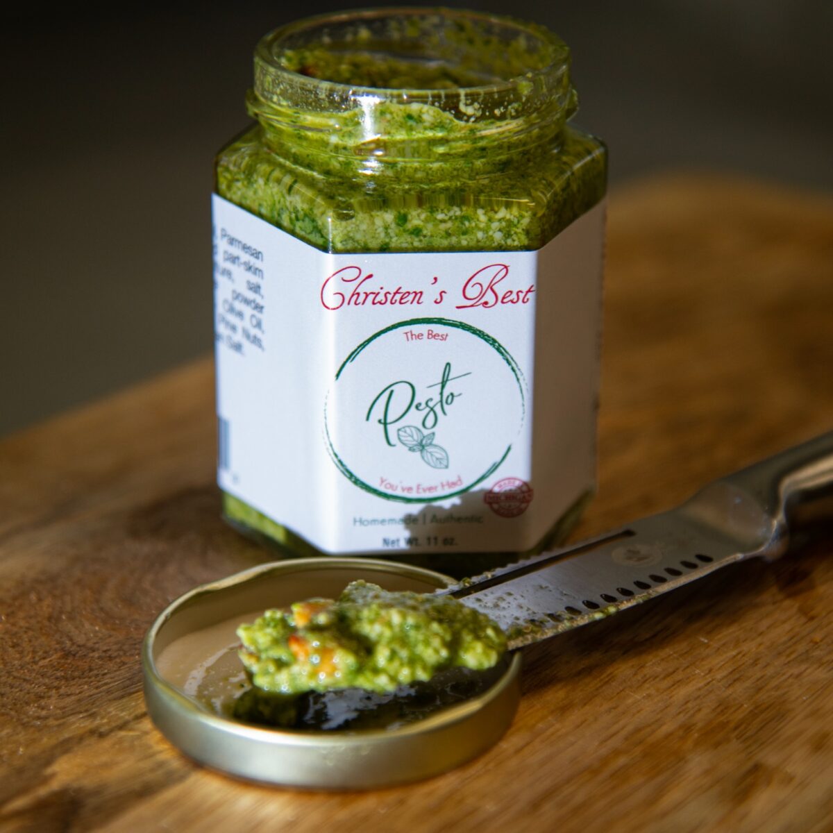 An open jar of Christen's Best pesto on a wooden board, with a spoonful resting on the lid beside a spreader