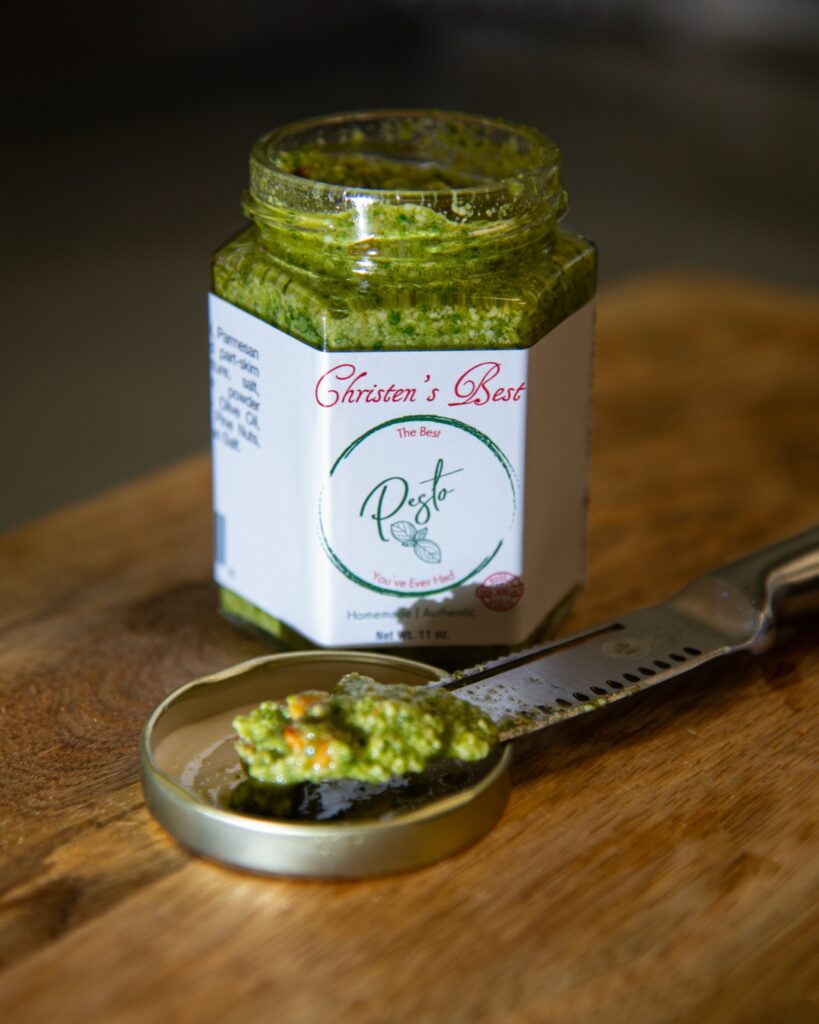 An open jar of Christen's Best pesto on a wooden board, with a spoonful resting on the lid beside a spreader