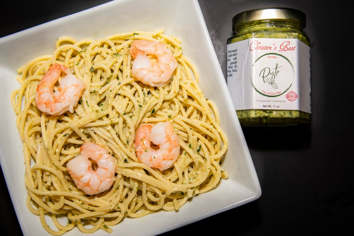 A square bowl of spaghetti tossed in basil pesto and topped with shrimp, beside a jar of Christen's Best