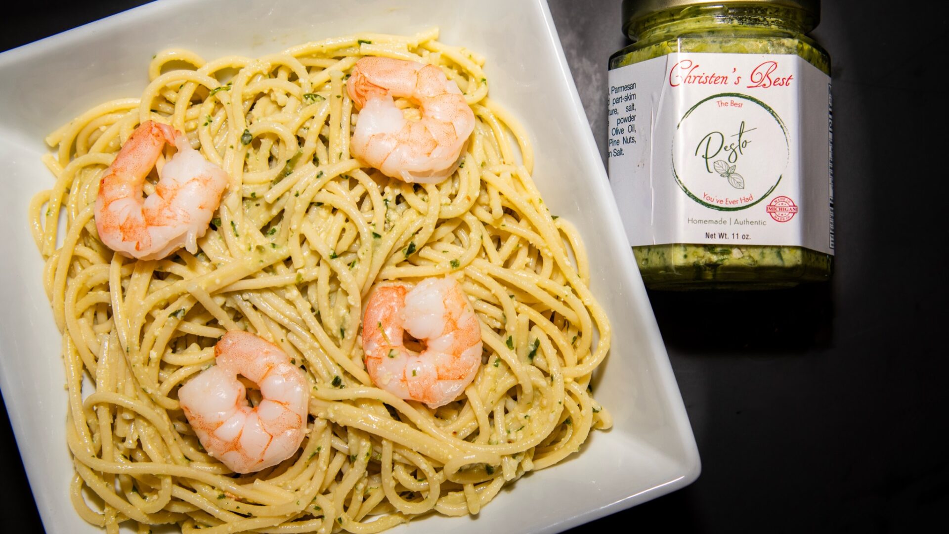 A square bowl of spaghetti tossed in basil pesto and topped with shrimp, beside a jar of Christen's Best
