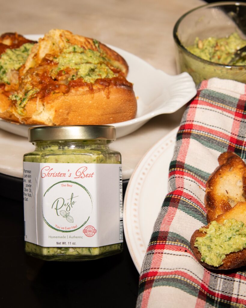 A jar of Christen's Best pesto in front of a toasted sub and a plate of pesto crostini on a plaid napkin